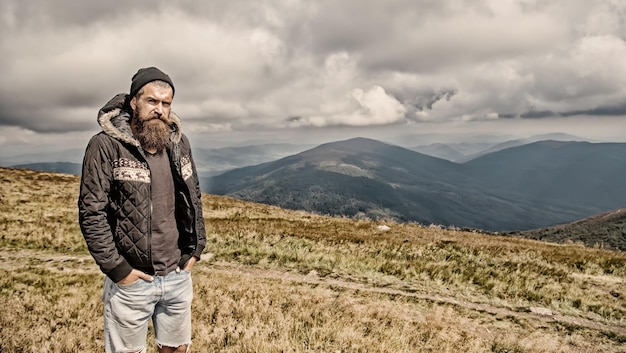 Hipster hombre barbudo barba larga brutal caucásico con bigote chico sin afeitar en chaqueta con cabello elegante obteniendo barbas corte de pelo cima de la montaña en el cielo nublado natural Hombre barbudo con barba larga