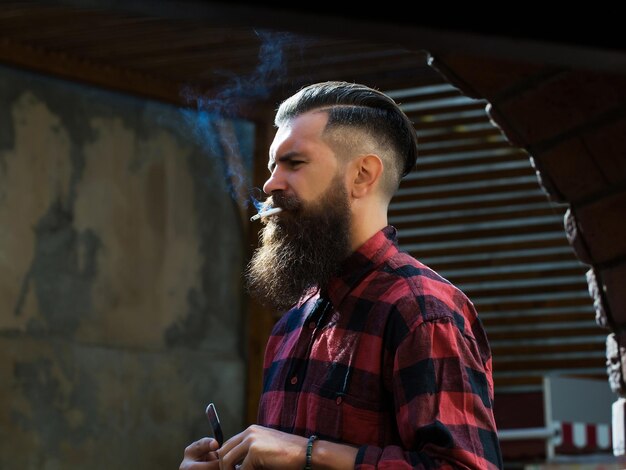 Hipster de hombre joven con cara guapo brutal barbudo con pelo de moda en camisa a cuadros roja y negra fumando cigarrillo en el fondo de arco de ladrillo interior