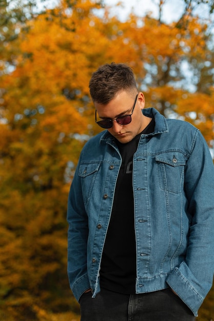 Hipster de hombre de otoño de moda en ropa casual con chaqueta de mezclilla y gafas de sol camina en el parque amarillo de otoño