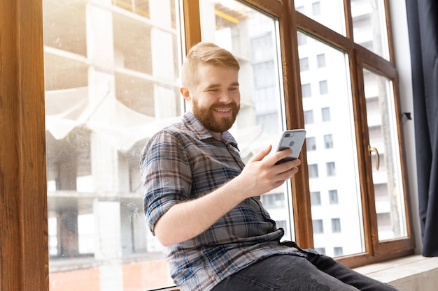 Hipster jovem bonito positivo com uma barba está sentado no parapeito da janela perto da grande janela e