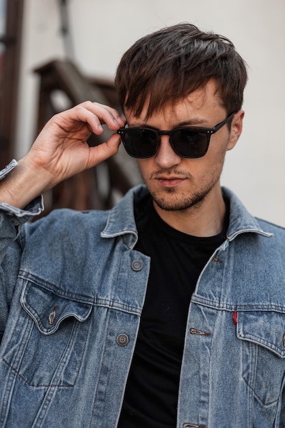 Hipster joven con estilo endereza las gafas de sol de moda. Retrato de un chico guapo con una chaqueta de mezclilla azul de moda en una camiseta negra al aire libre en la ciudad. Estilo americano.