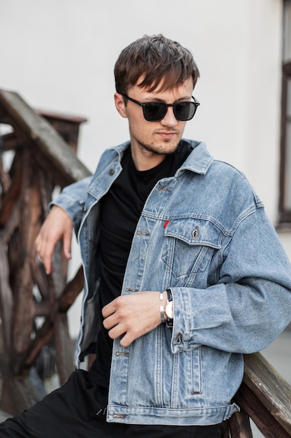 Hipster joven guapo con una chaqueta de mezclilla azul de moda en elegantes gafas de sol en jeans negros está descansando en la ciudad cerca del edificio vintage. Chico atractivo se relaja al aire libre en la ciudad. Ropa de caballero.