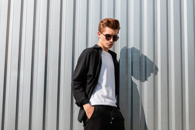 Hipster joven moderno con una elegante camisa en una camiseta con pantalones a rayas de moda en gafas de sol cerca de la pared de metal plateado en la ciudad. Buen chico con un peinado elegante se relaja al aire libre en un día soleado.