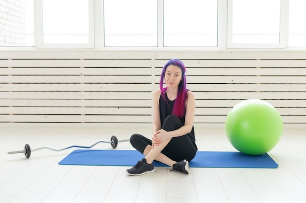 Hipster joven de raza mixta con cabello morado está sentado en la colchoneta de deporte junto a la barra y con el fitball en el gimnasio. Concepto de fitness y ejercicio regular.