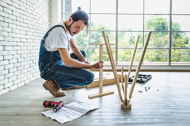 Hipster junger Mann, der als Handwerker arbeitet, der beim Zusammenbauen von Holztischen mit Ausrüstungen, Konzept für Heimwerken und Selbstbedienung nach Handbuch sucht.