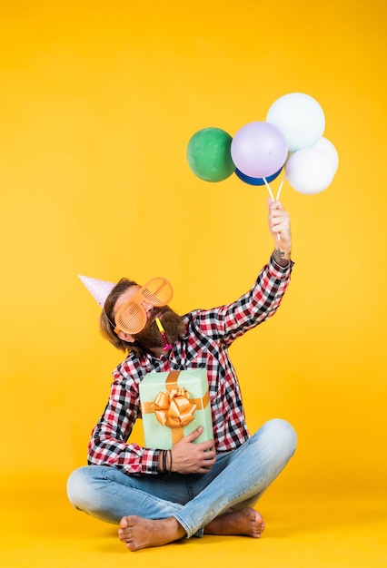 Hipster maduro caucásico con gafas de fiesta divertidas en camisa a cuadros sosteniendo globos y caja de regalo, celebración.