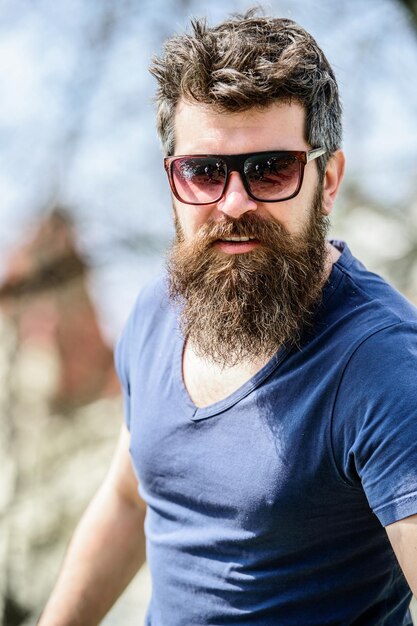 Homem barbudo bonito americano usando óculos escuros, boné e jaqueta de  capuz contra o céu azul. retrato de cara brutal russo caucasiano parece  para o lado, pose relaxada com rosto natural. copie