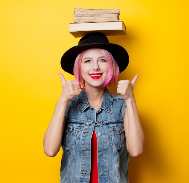 Hipster-Mädchen im jungen Stil mit rosafarbener Frisur, die Bücher über dem Kopf auf gelbem Hintergrund hält