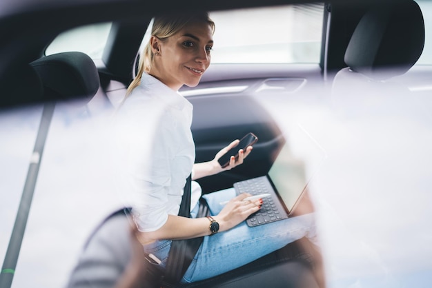 Hipster-Mädchen mit Handy und tragbarem PC, das auf dem Weg zum Geschäftstreffen wegschaut und lächelt