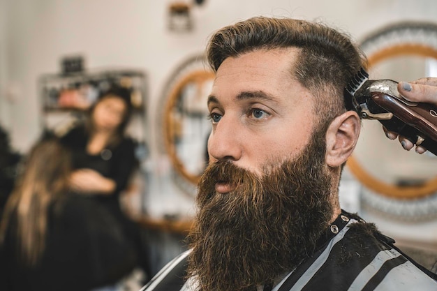 Hipster-Mann im Barbershop für Bart- und Haarschneiden Weibliche Hände der Friseurfrau mit Haarschneidemaschine für den klassischen Gentleman-Schnitt Friseursalon-Konzept