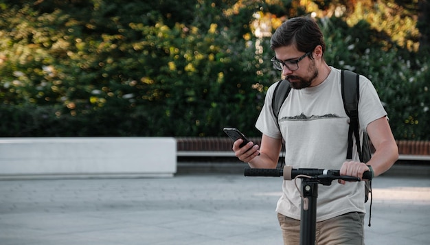 Hipster-Mann-Pendler mit Elektroroller im Freien in der Stadt mit Smartphone