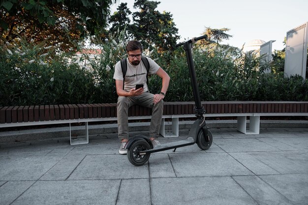 Hipster-Mann-Pendler mit Elektroroller im Freien in der Stadt mit Smartphone
