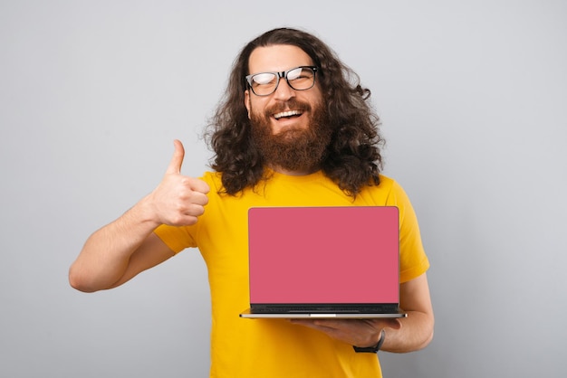 Hipster-Mann zeigt Daumen nach oben und hält einen Laptop mit leerem Bildschirm Studioaufnahme über grauem Bakcground
