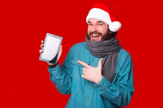 Hipster masculino alegre usando um chapéu de Natal está apontando para um tablet.