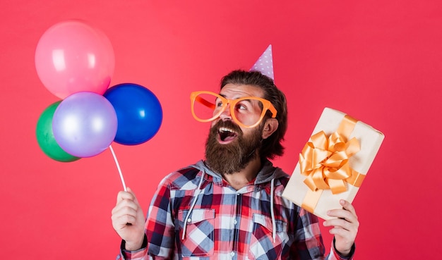 Foto hipster masculino con barba y cabello peinado a la moda sostiene globos de fiesta y caja de cumpleaños