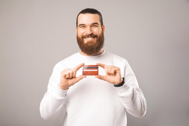 Hipster masculino barbudo alegre está sosteniendo una tarjeta de crédito con ambas manos