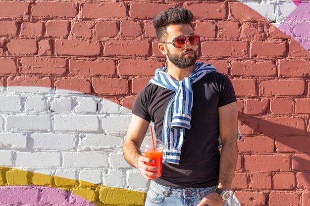 Hipster masculino joven árabe elegante lindo con un bigote y una barba y con gafas bebiendo jugo con una pajita. Concepto de vacaciones de verano.