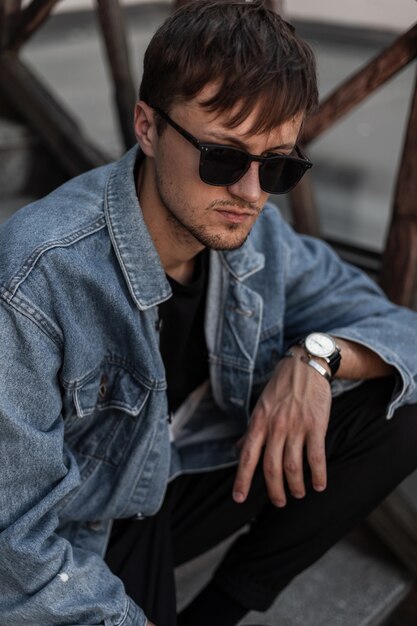 Hipster de moda joven en ropa de mezclilla con estilo moderno en gafas de sol negras se sienta en una escalera vintage en la ciudad en un día de primavera. Chico atractivo atractivo descansando al aire libre. Moda masculina.