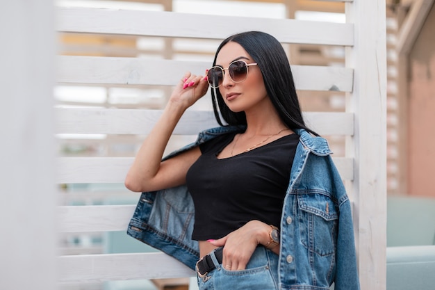 Hipster de mujer joven con estilo urbano con pelo largo negro en un top negro en una chaqueta de mezclilla de moda en una falda con gafas de sol posando junto a una pared de madera blanca en la ciudad. Modelo de moda chica atractiva.