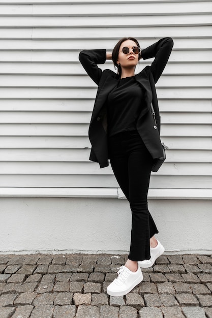 Hipster de mujer joven de moda en gafas de sol en ropa negra juvenil de moda en zapatillas de deporte blancas de moda con bolso de cuero se encuentra cerca de la pared vintage de madera al aire libre. Modelo de moda chica camina en la calle.