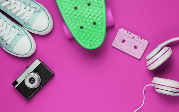 Hipster-Outfit. Skateboard, Kopfhörer, Audiokassette, Turnschuhe, Retro-Kamera auf rosa Hintergrund. Kreativer Mode-Minimalismus. Minimaler Sommerspaß. Pop-Art. 80er Jahre. Platz kopieren. Ansicht von oben