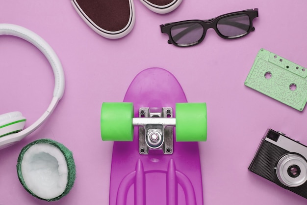 Hipster-Outfit. Skateboard mit Kopfhörern, Audiokassette, Retro-Kamera und Turnschuhen auf rosa Hintergrund. Kreativer Mode-Minimalismus. Minimaler Sommerspaß. Pop-Art. 80er Jahre.