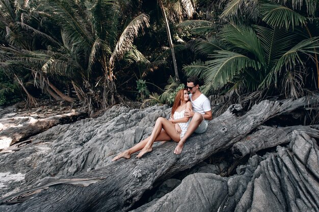 Hipster-Paar am Strand, Mädchen mit roten Haaren im weißen Kleid und Mann mit Stoppeln, die auf dem alten großen kahlen Baum ohne Blätter sitzen. Phuket. Thailand