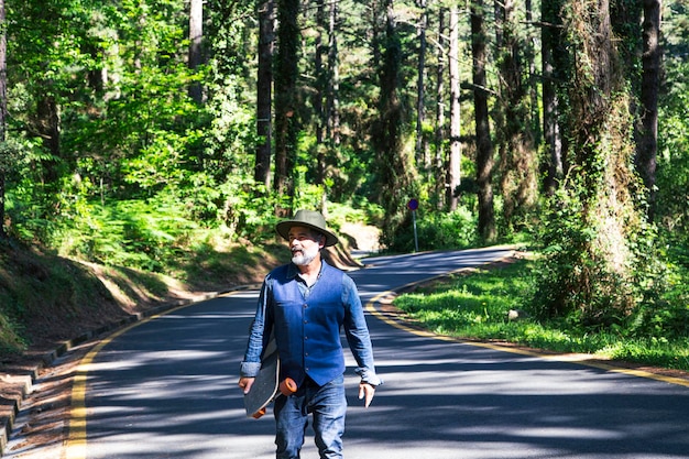 Hipster con una patineta longboard en sus manos camina por un camino en el bosque