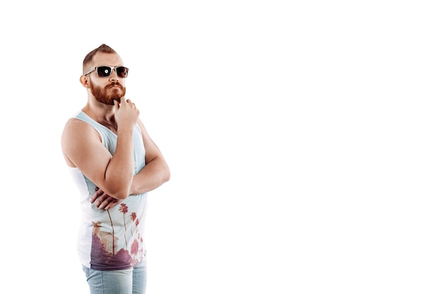 Hipster pelirrojo hombre barbudo en gafas de sol en verano posando mientras está de pie sobre fondo blanco.