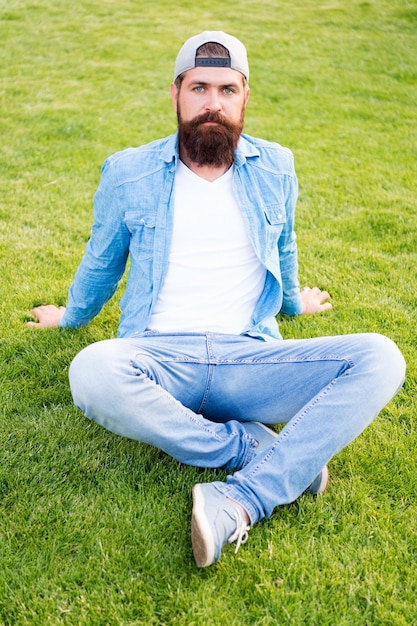 Hipster peludo. Hipster brutal sentado en la hierba verde. Hombre barbudo en estilo hipster de moda en verano al aire libre. Hipster caucásico con pelo grueso de barba con gorra de béisbol.