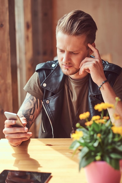 Hipster pensativo com um corte de cabelo elegante e barba se senta em uma mesa em um café na estrada, usando um smartphone.