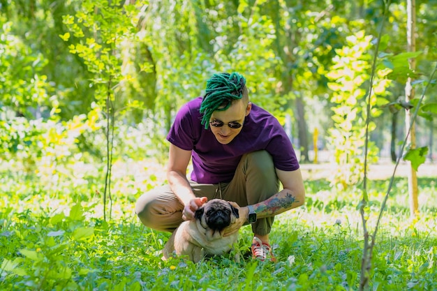 Un hipster con rastas verdes y gafas redondas negras está sentado en un césped verde y acariciando a un pug lindo en un día soleado de verano Un hombre camina un pug