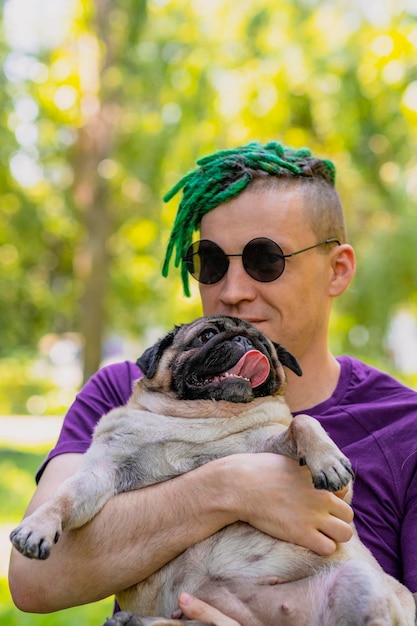 Hipster con rastas verdes y gafas redondas negras sosteniendo y acariciando a un pug lindo en un día soleado de verano Un hombre pasea a su perro