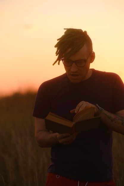 Hipster sorridente com livro Ângulo baixo de jovem macho com dreadlocks em roupas casuais e óculos em pé com livro contra o céu pôr do sol