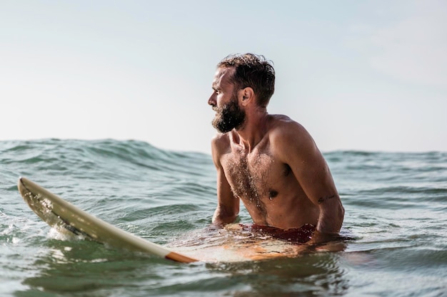 Hipster surfista sentado en su tabla de surf en el agua del océano y esperando una gran ola Hombre barbudo en forma entrenando con tabla de surf al mar Concepto de aventura y libertad haciendo deportes acuáticos extremos