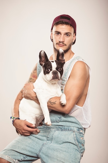 Hipster-Typ, der seinen netten Hund der französischen Bulldogge vor neutralem Hintergrund umarmt