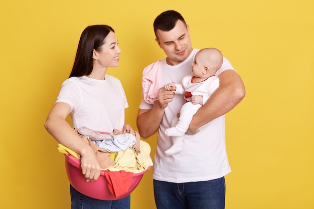 Hipster Vater und Mutter posieren mit ihrem Kind isoliert über gelbe Wand, attraktive Frau, die Becken mit Kleidung hält, Wäsche macht, niedliches Baby auf Papas Händen.