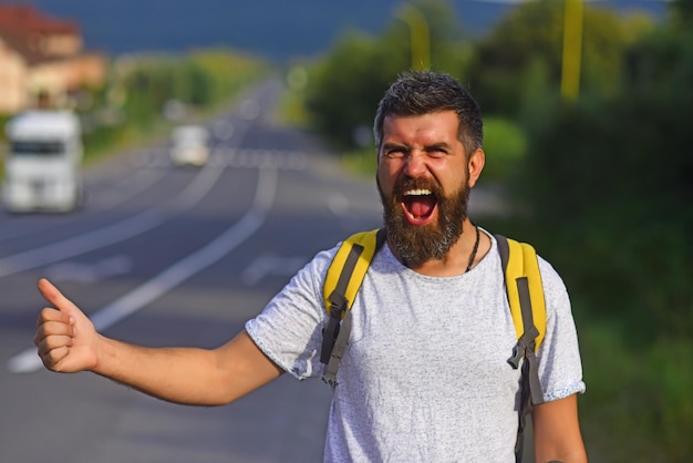 Hipster de viaje de parada automática intenta detener el coche y el pulgar hacia arriba gesto hombre con cara alegre y barba trave