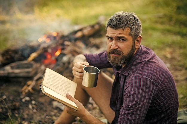 Hipster-Wanderer mit Buch und Becher am Lagerfeuer über die Natur