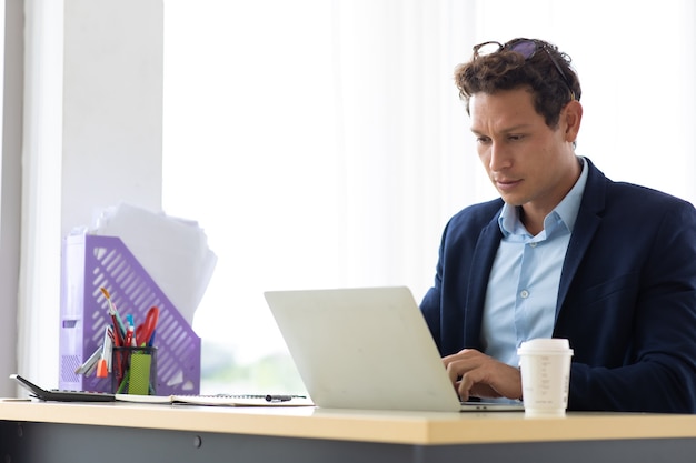 Hispânico jovem sentado beber café e trabalhando no computador portátil no escritório em casa criativo.