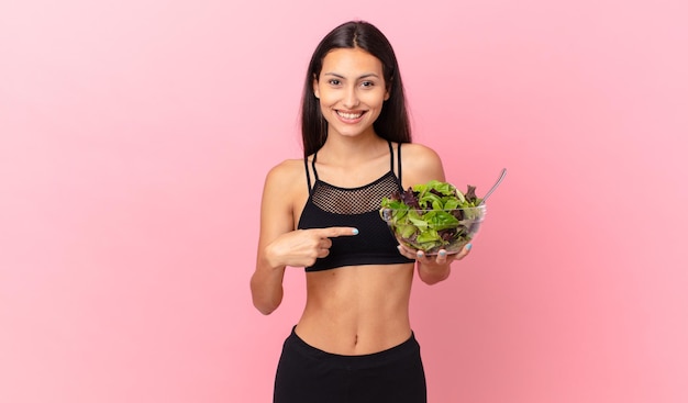 Hispanische Fitnessfrau, die aufgeregt und überrascht aussieht, auf die Seite zeigt und einen Salat hält