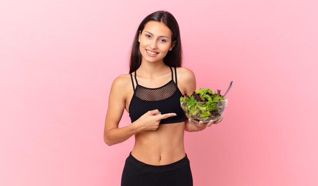 Hispanische Fitnessfrau, die fröhlich lächelt, sich glücklich fühlt und auf die Seite zeigt und einen Salat hält