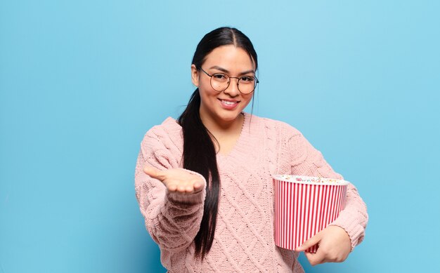 Hispanische Frau, die glücklich mit freundlichem, selbstbewusstem, positivem Blick lächelt und ein Objekt oder Konzept anbietet und zeigt