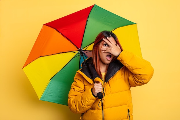 Hispanische Frau, die schockiert, verängstigt oder verängstigt aussieht und das Gesicht mit der Hand bedeckt. Regen- und Regenschirmkonzept