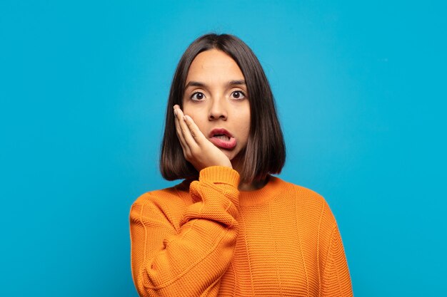 Hispanische Frau, die sich schockiert und erstaunt fühlt, Gesicht zu Hand ungläubig mit weit geöffnetem Mund haltend