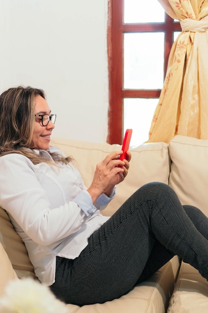 Hispanische Frau, die zu Hause auf der Couch liegt Sie hält ein Smartphone in der Hand, das sie ansieht und lächelt