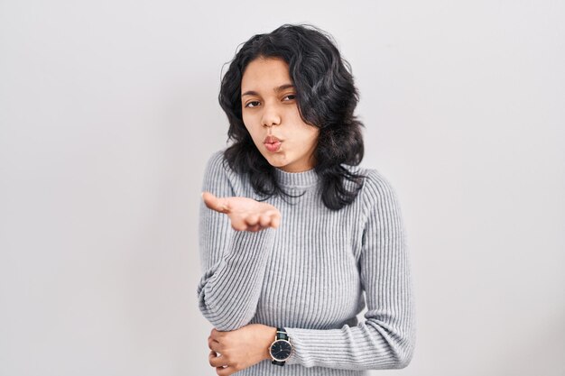 Hispanische Frau mit dunklem Haar, die vor isoliertem Hintergrund steht und in die Kamera blickt und einen Kuss mit der Hand in die Luft bläst, was ein schöner und sexy Liebesausdruck ist