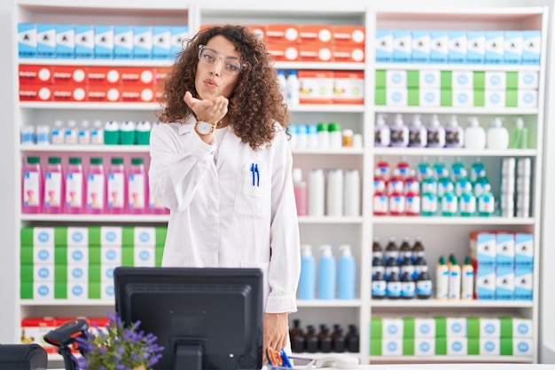 Hispanische Frau mit lockigem Haar, die in einer Apotheke arbeitet und in die Kamera schaut und einen Kuss mit der Hand auf die Luft bläst, ist schön und sexy. Liebe Ausdruck.