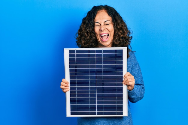 Hispanische Frau mittleren Alters, die ein Photovoltaik-Solarpanel hält, lächelt und lacht laut, weil lustiger, verrückter Witz.