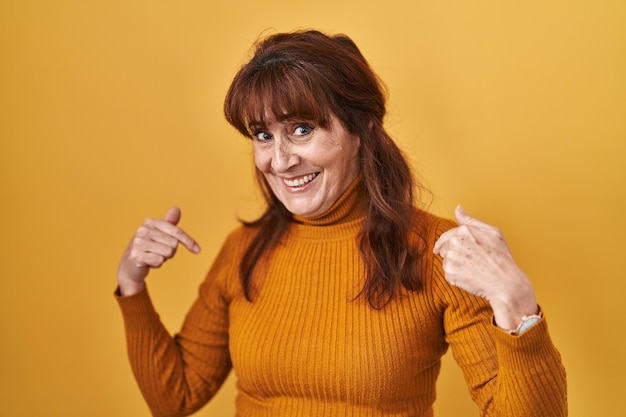 Hispanische Frau mittleren Alters, die über gelbem Hintergrund steht und selbstbewusst mit einem Lächeln im Gesicht aussieht und sich mit stolzen und glücklichen Fingern zeigt.